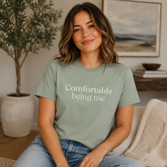 Woman wearing a Sage Comfortable Being Me T-Shirt with cream serif text, seated in a cosy neutral living room.