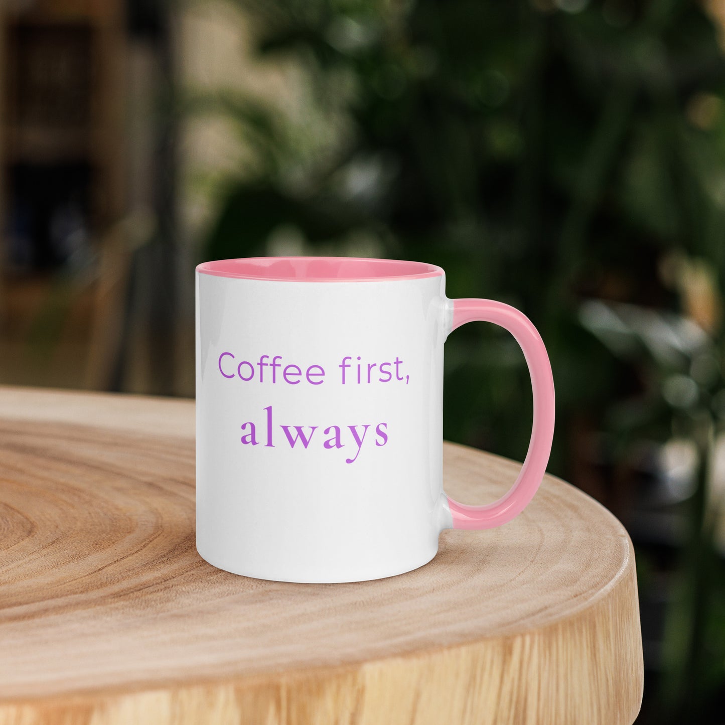 Ceramic coffee mug with pink handle and the phrase “coffee first, always”, placed on a wooden table with warm tones and soft lighting for a calm and inviting coffee moment.