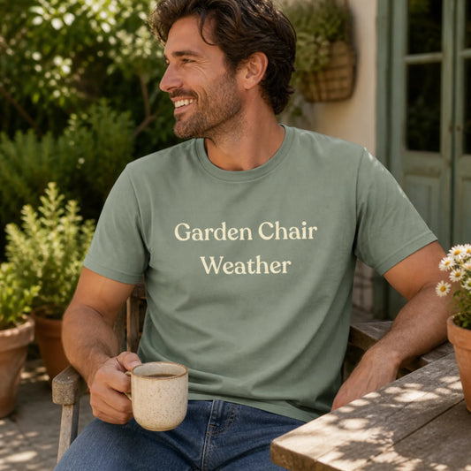 Male model wearing the Garden Chair Weather T-Shirt in Sage, sitting outside with a mug in a relaxed garden patio setting.