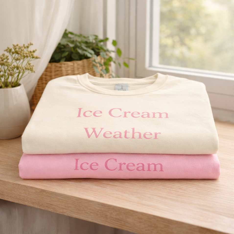 Folded Ice Cream Weather T-shirts in ivory and blossom stacked on wooden shelf, soft cotton summer lifestyle tee