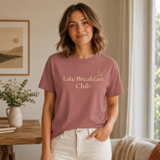 Female model wearing a mauve Late Breakfast Club T-shirt with soft yellow text in a warm relaxed kitchen setting.