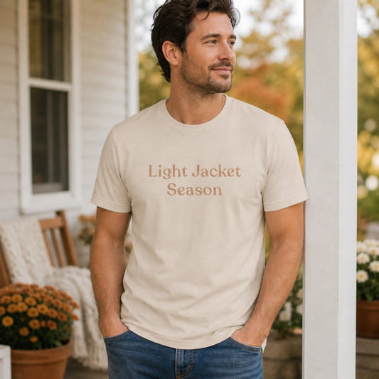 Man wearing the Light Jacket Season T-shirt in soft cream with warm muted text, standing outdoors on a porch in soft seasonal light.