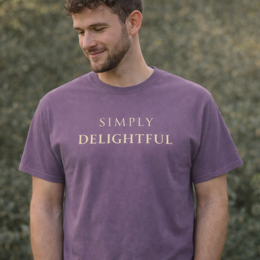 Man wearing muted purple “Simply Delightful” graphic T-shirt outdoors in warm natural light with a relaxed, content expression