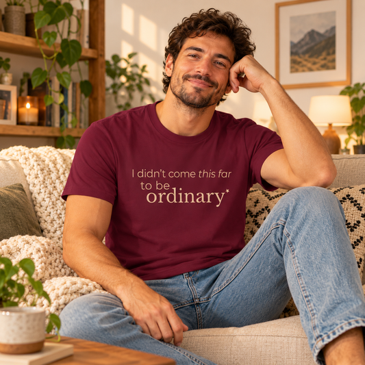 Man wearing a maroon “I Didn’t Come This Far to Be Ordinary” T-shirt while sitting casually on a sofa in a warm, cosy living room.