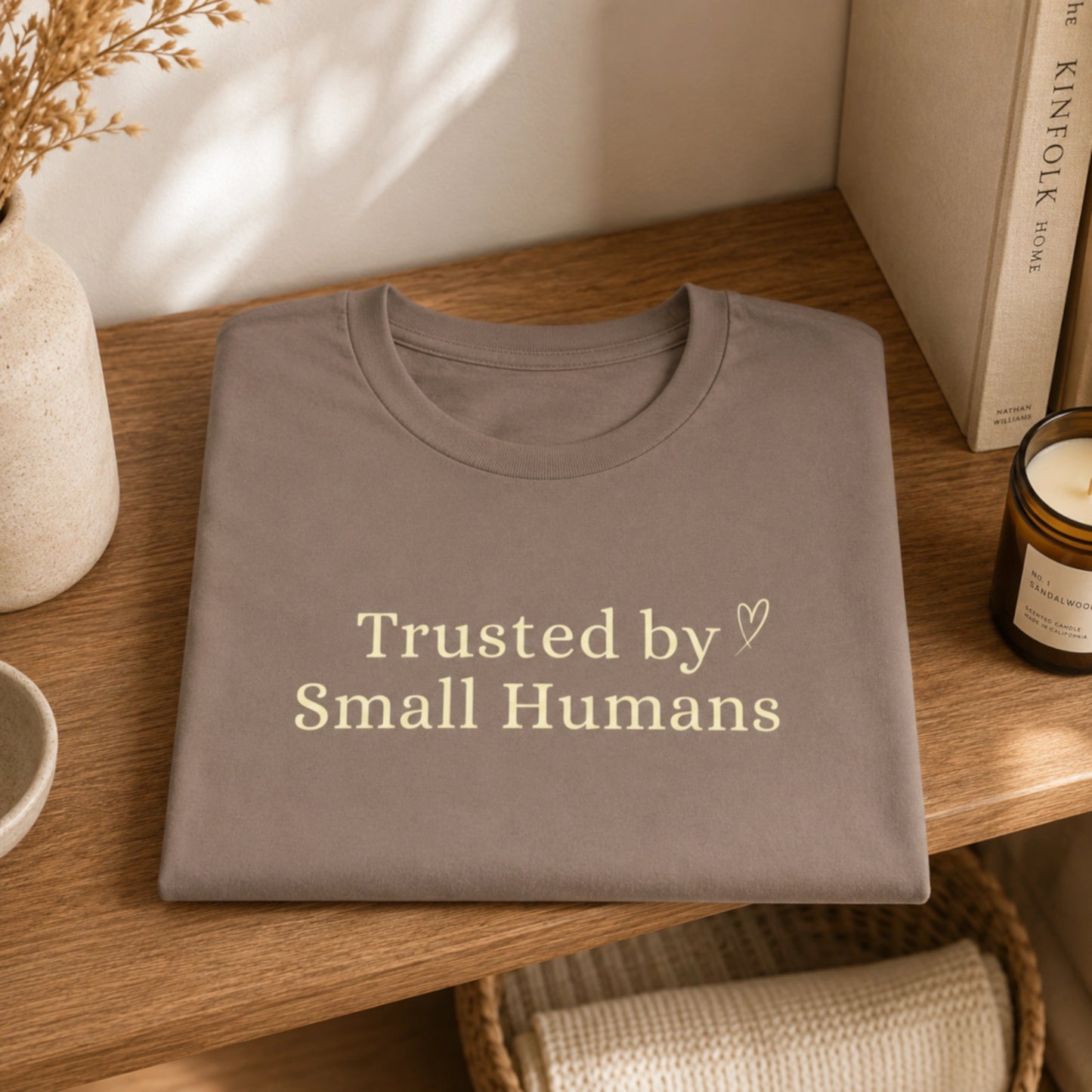 Folded Trusted by Small Humans T-Shirt in Pebble on a wooden shelf, featuring soft cream serif lettering and a small heart detail.