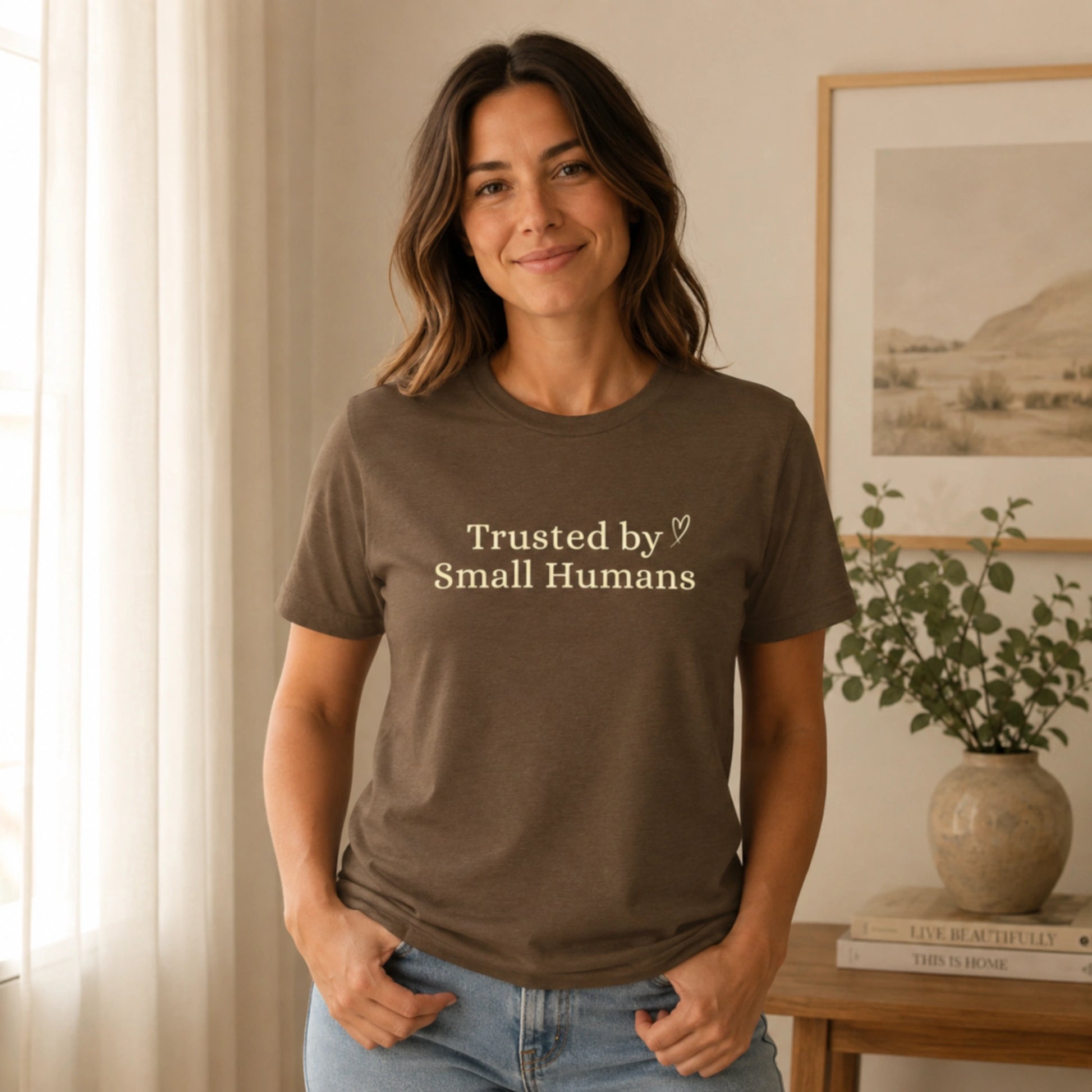 Female model wearing the Trusted by Small Humans T-Shirt in Heather Brown, featuring soft cream serif lettering and a small heart detail on a Bella+Canvas unisex tee.