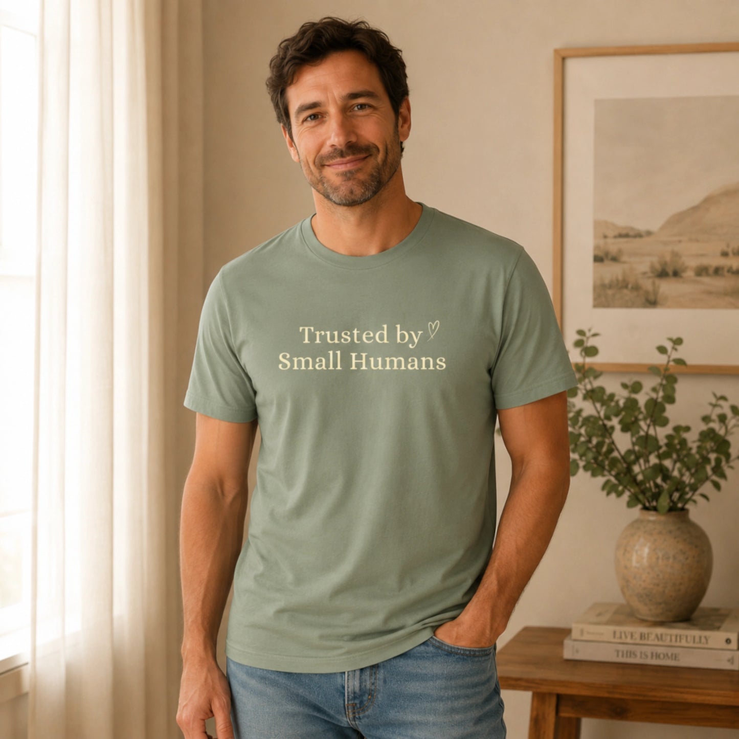 Male model wearing the Trusted by Small Humans T-Shirt in Sage, featuring soft cream serif lettering and a small heart detail on a Bella+Canvas unisex tee.
