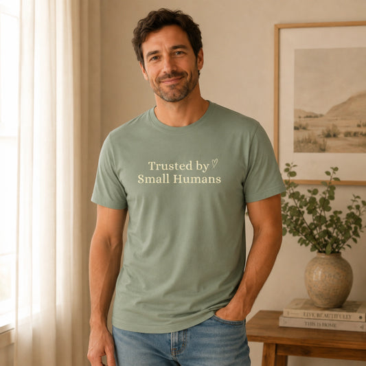 Male model wearing the Trusted by Small Humans T-Shirt in Sage, featuring soft cream serif lettering and a small heart detail on a Bella+Canvas unisex tee.