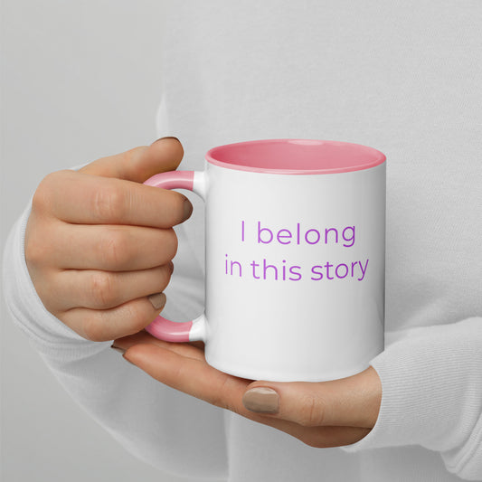 Ceramic coffee mug with pink handle and the words “I belong in this story”, held in hands with soft knit sleeves, capturing a cosy and imaginative reading moment.