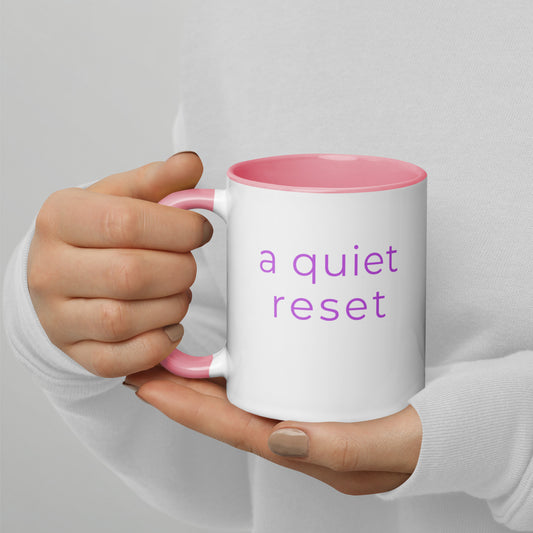Ceramic coffee mug with pink handle and the words “a quiet reset”, held in hands with soft knit sleeves, capturing a peaceful and reflective moment.