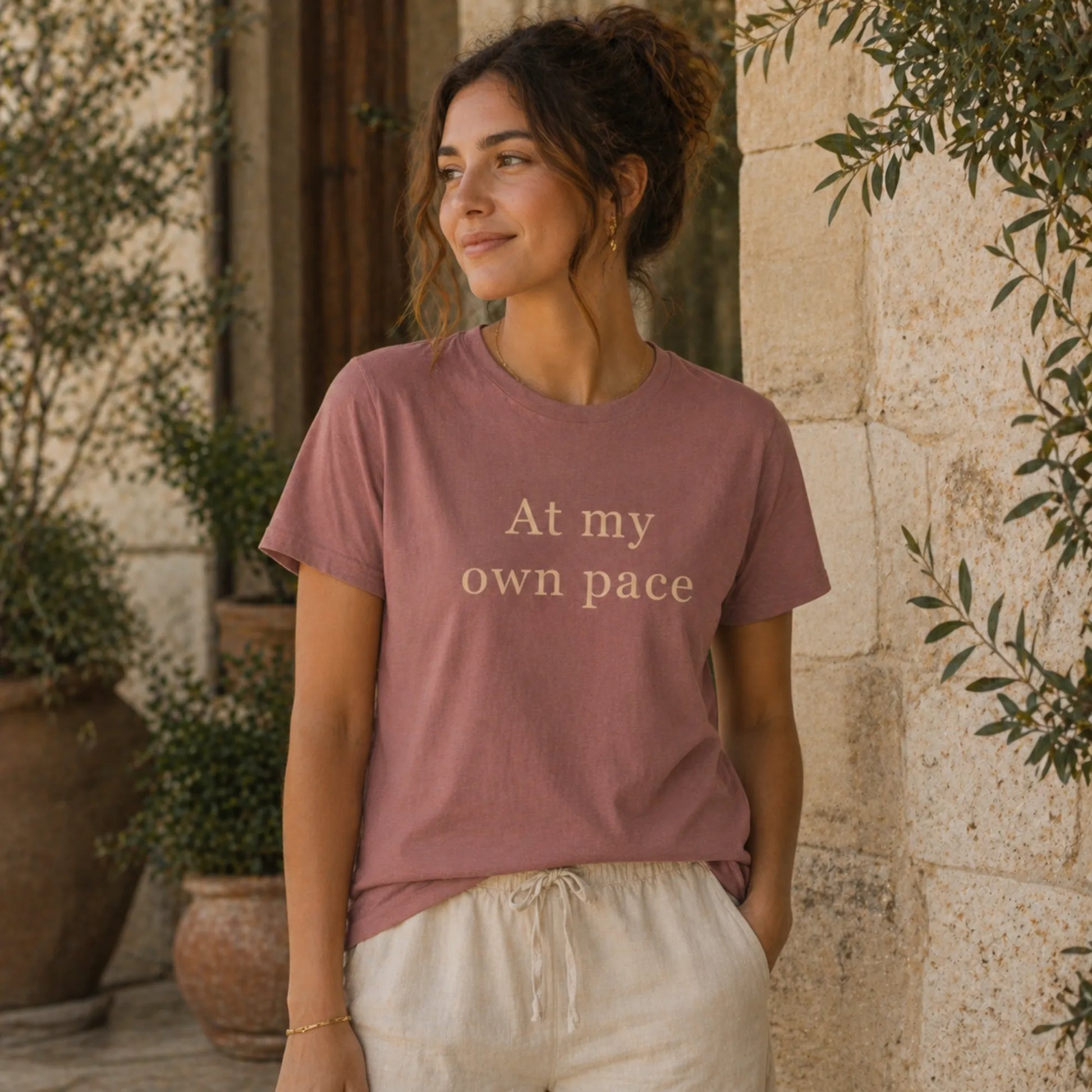 Female model wearing an At My Own Pace T-Shirt in heather mauve in a calm, natural lifestyle setting.
