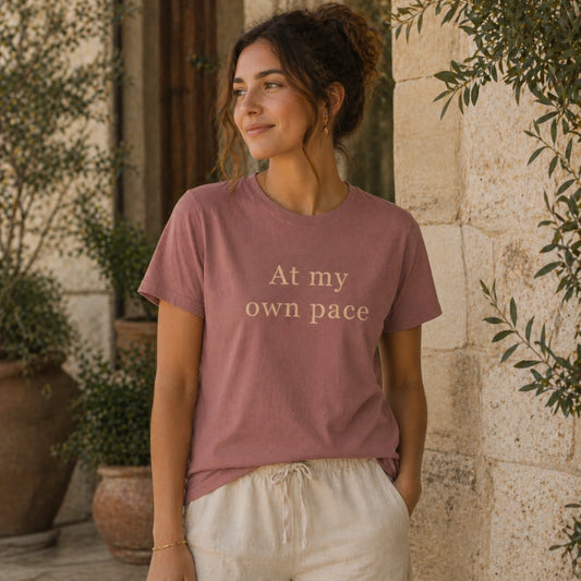 Female model wearing an At My Own Pace T-Shirt in heather mauve in a calm, natural lifestyle setting.