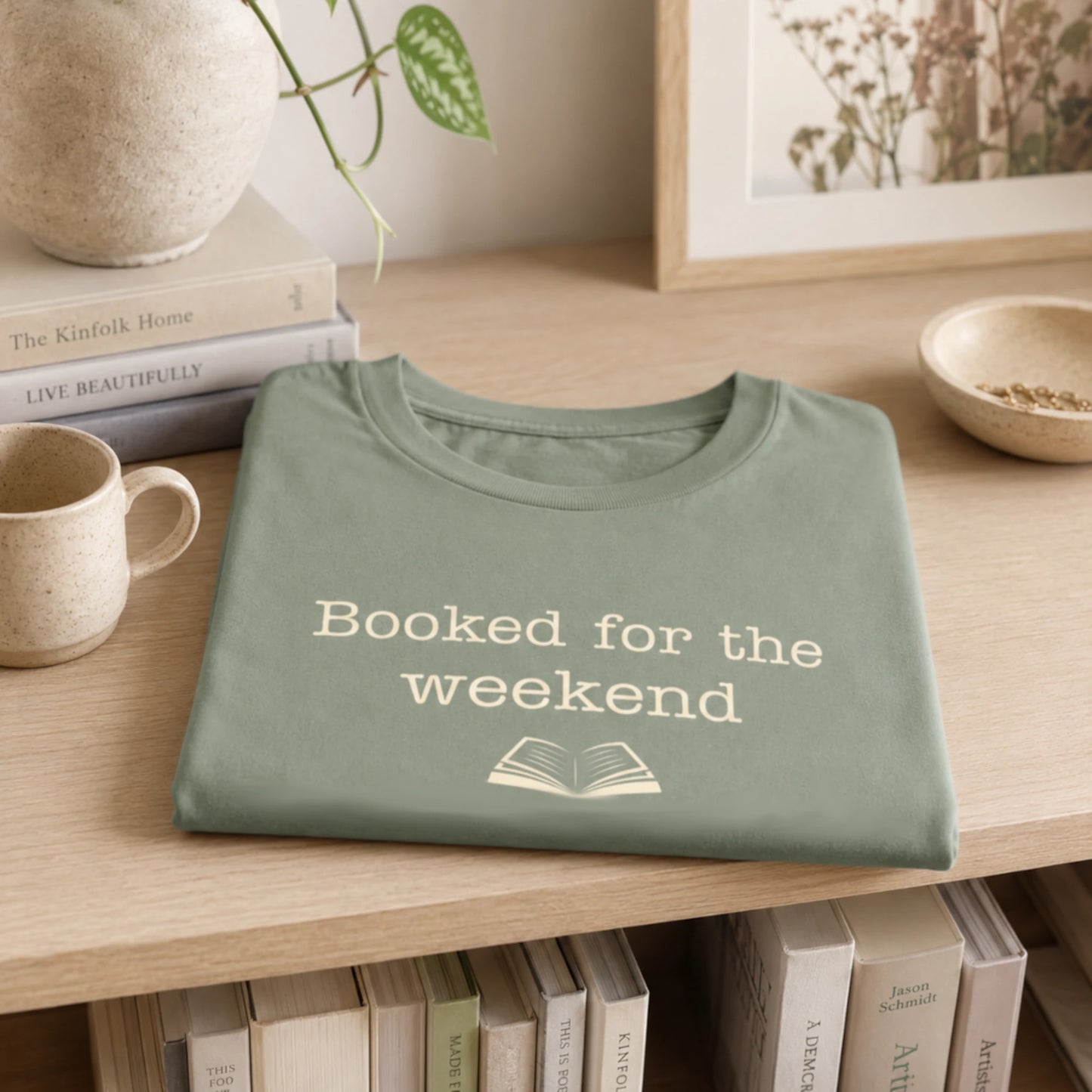 Folded sage “Booked for the Weekend” T-Shirt styled on a soft neutral shelf with books, a mug, and simple cosy home décor.