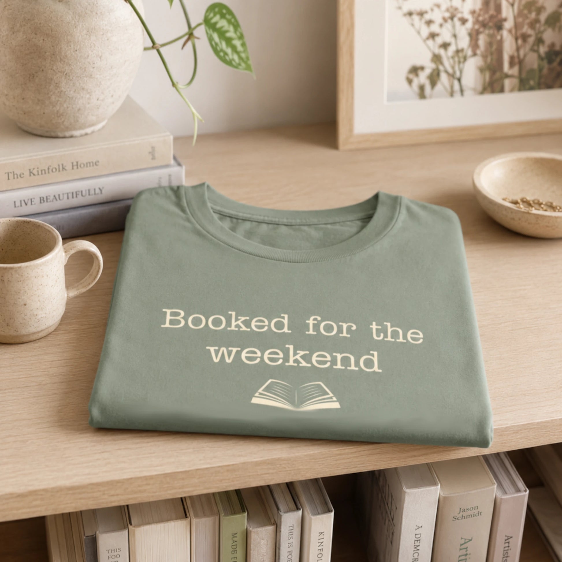 Folded sage “Booked for the Weekend” T-Shirt styled on a soft neutral shelf with books, a mug, and simple cosy home décor.