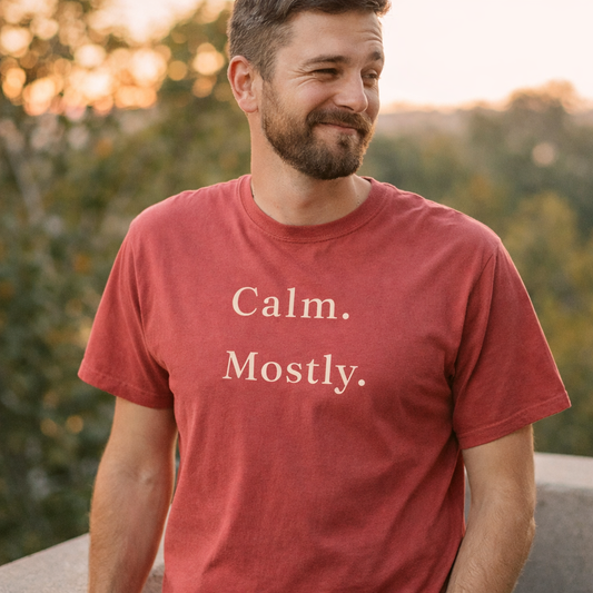 Man wearing crimson Comfort Colors T-shirt with “Calm. Mostly.” text, relaxed outdoor setting in warm evening light