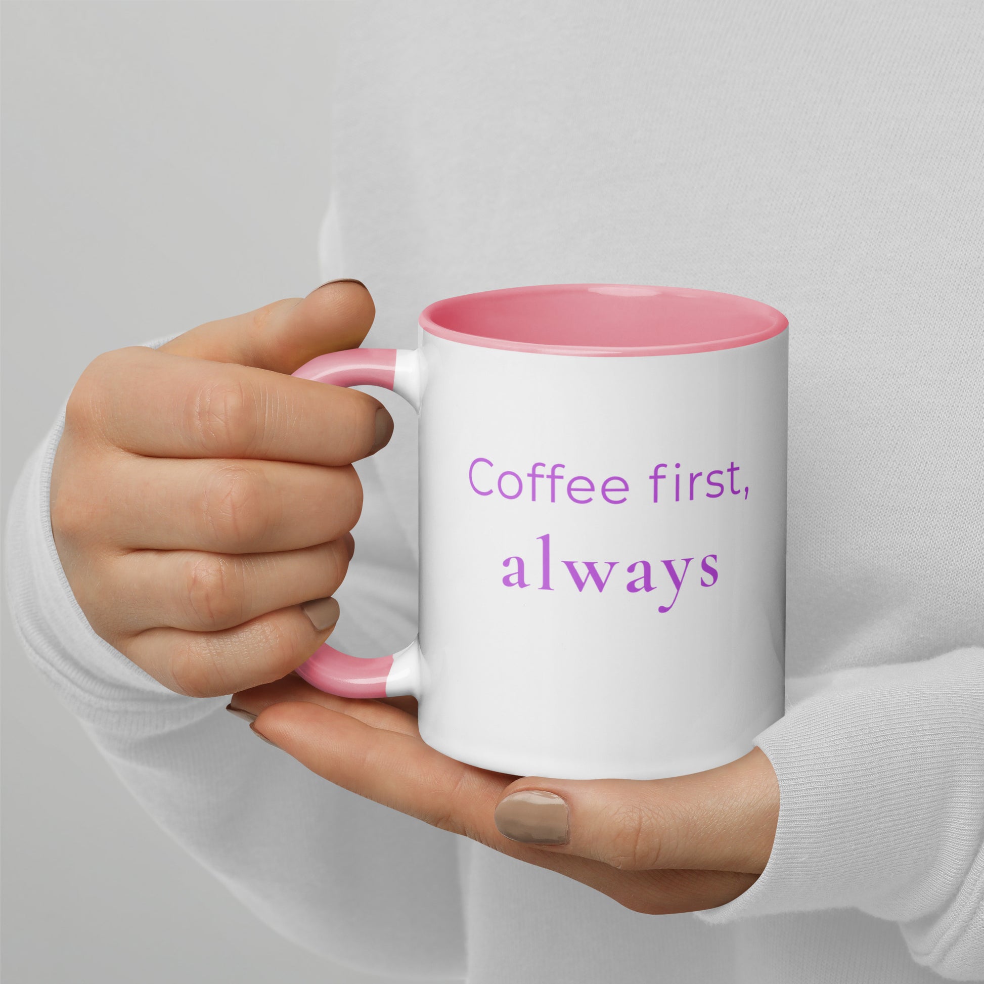 Ceramic coffee mug with pink handle and the words “coffee first, always”, held in hands with soft knit sleeves, capturing a warm and familiar coffee ritual.