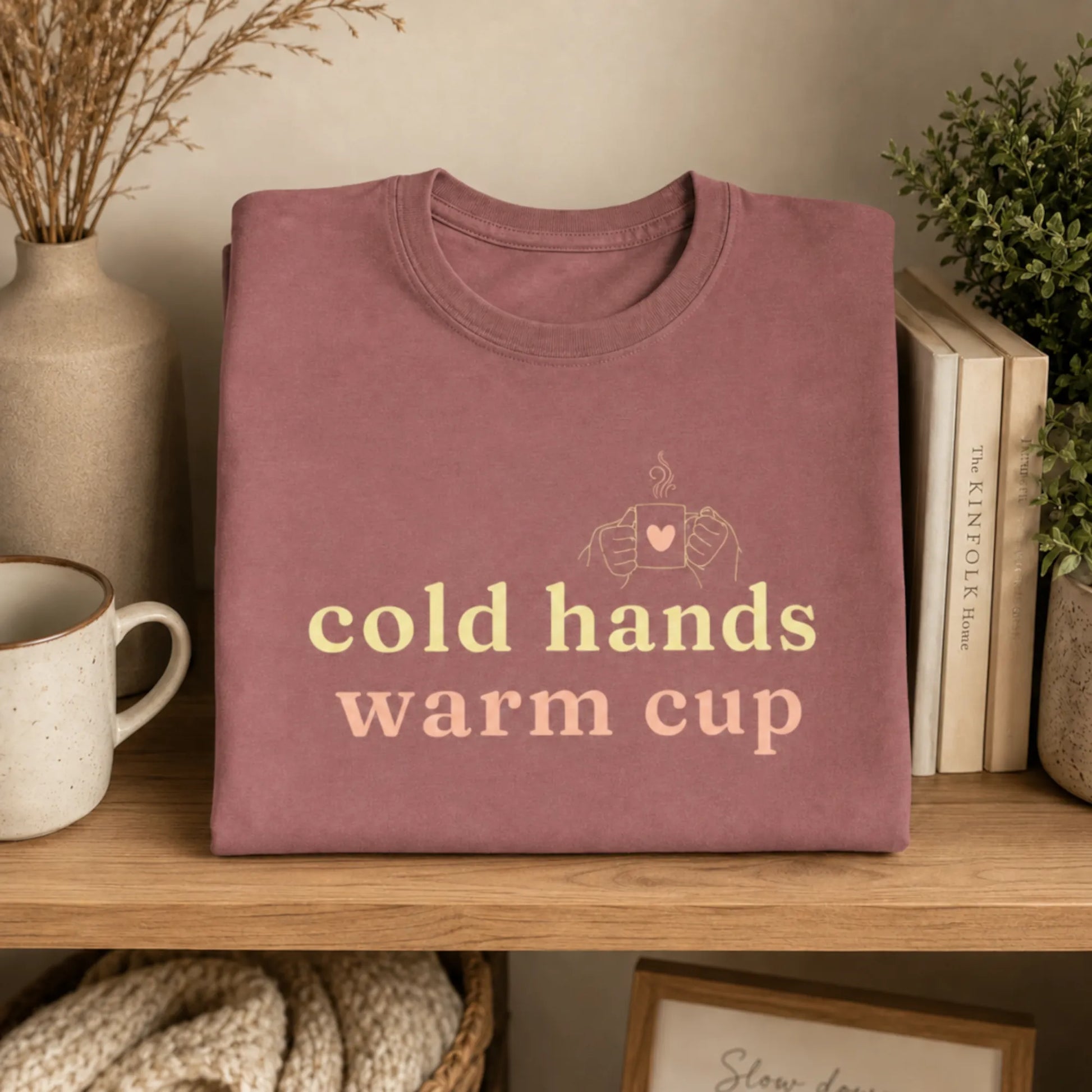 Folded heather mauve Cold Hands Warm Cup T-Shirt displayed on a wooden shelf with a ceramic mug, books, dried stems and cosy neutral home decor.