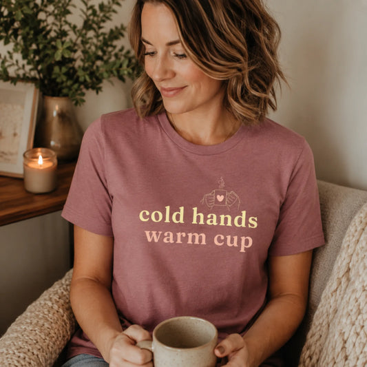 Female model wearing a heather mauve Cold Hands Warm Cup T-shirt, styled in a cosy setting with soft warm tones and a relaxed everyday feel.