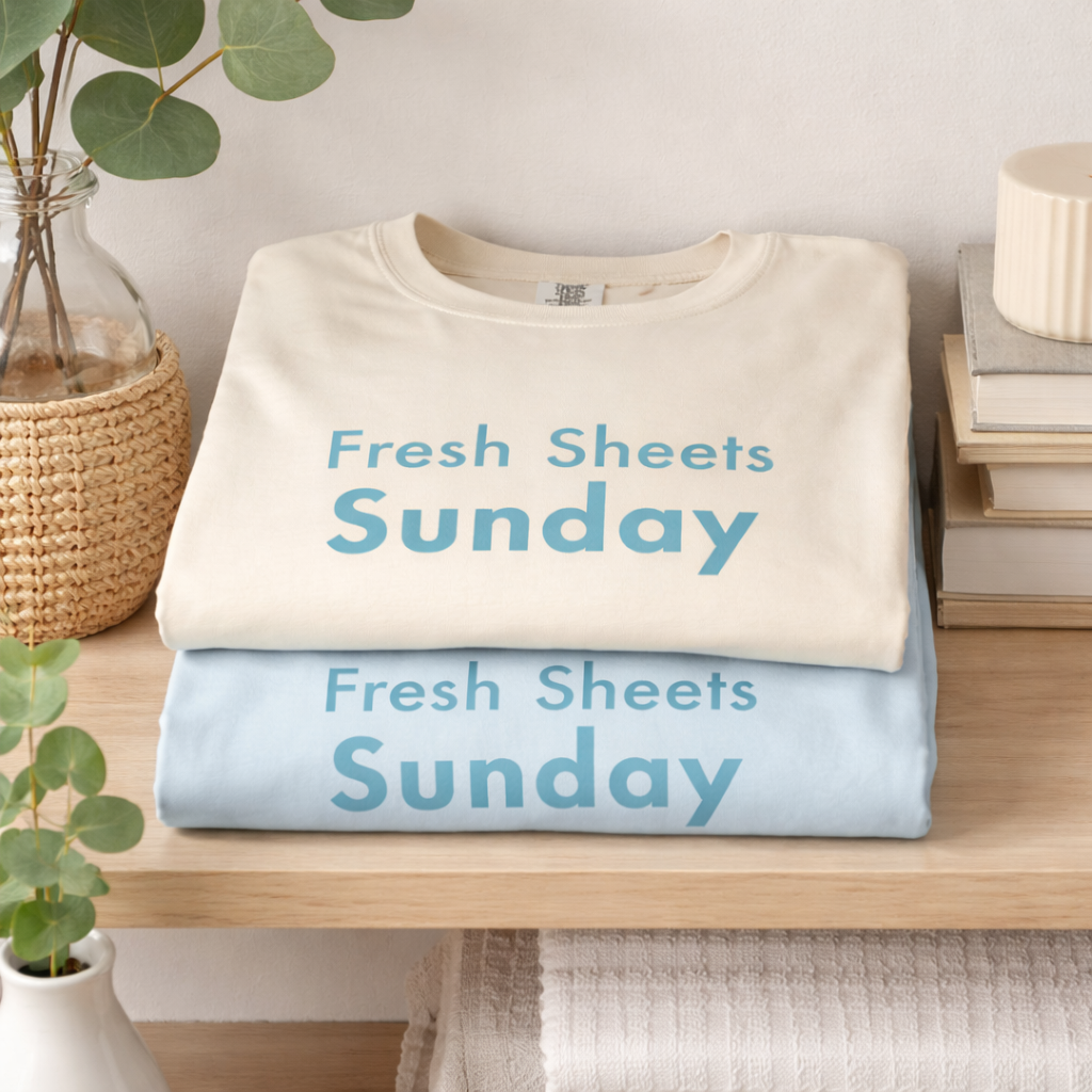 Folded Fresh Sheets Sunday Comfort Colors T-shirts in ivory and chambray styled on wooden shelf with soft home decor