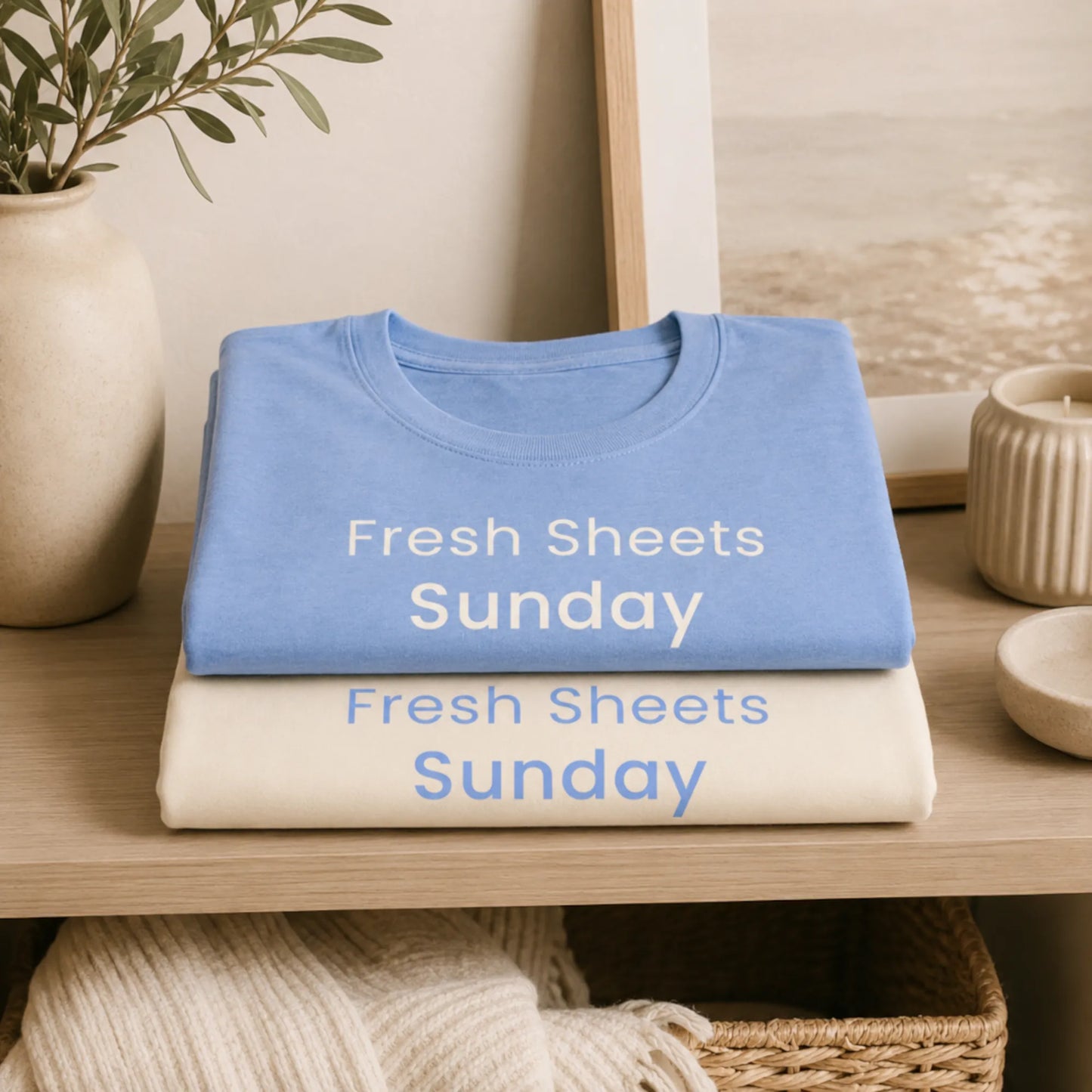 Folded Fresh Sheets Sunday T-shirts in natural and heather carolina blue displayed on a cosy home shelf with soft neutral decor.