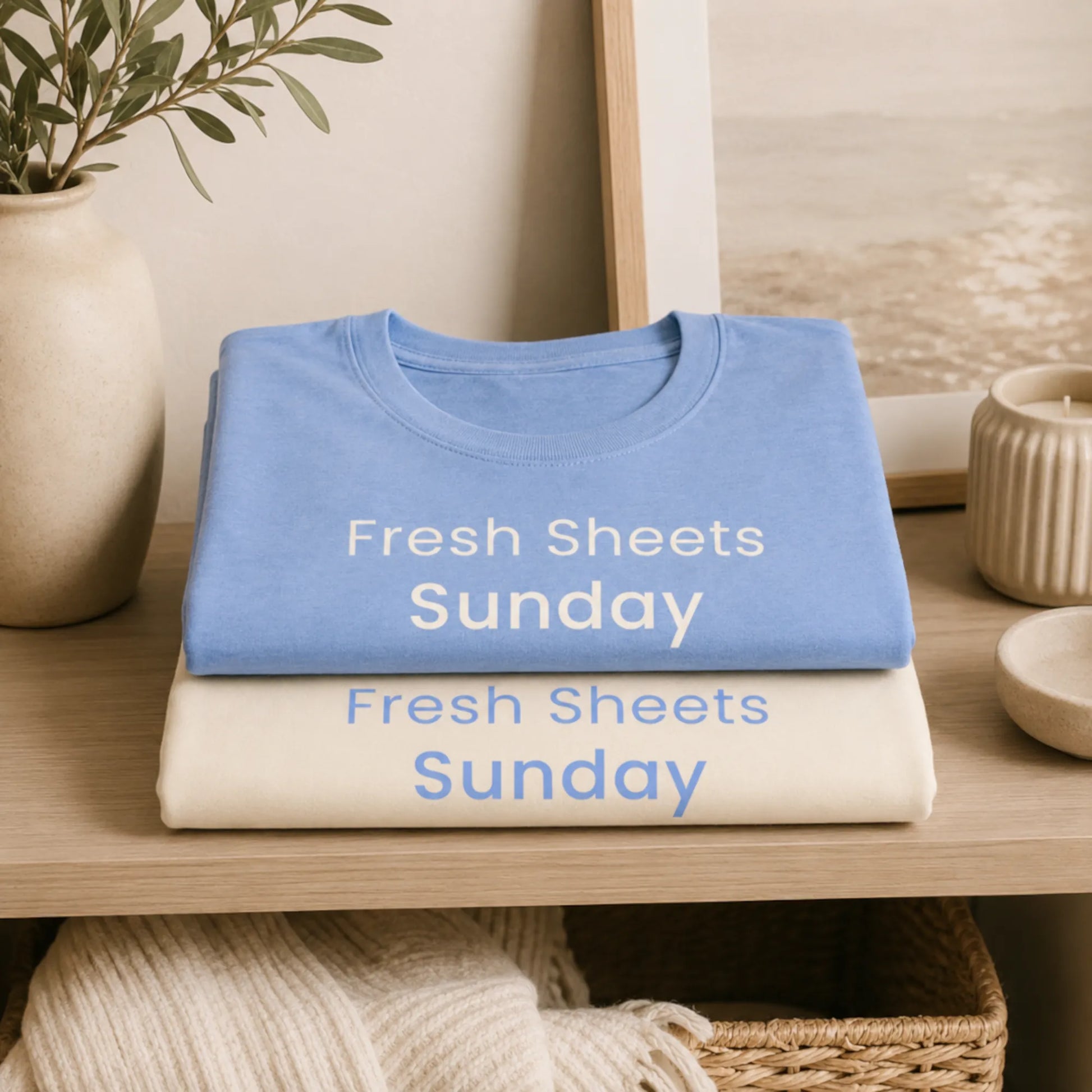 Folded Fresh Sheets Sunday T-shirts in natural and heather carolina blue displayed on a cosy home shelf with soft neutral decor.