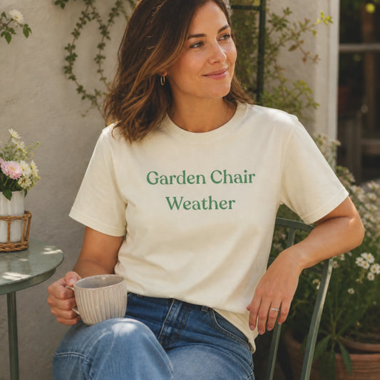 Female model wearing the Garden Chair Weather T-Shirt in Natural, sitting outdoors in a sunny garden setting with flowers and a mug.
