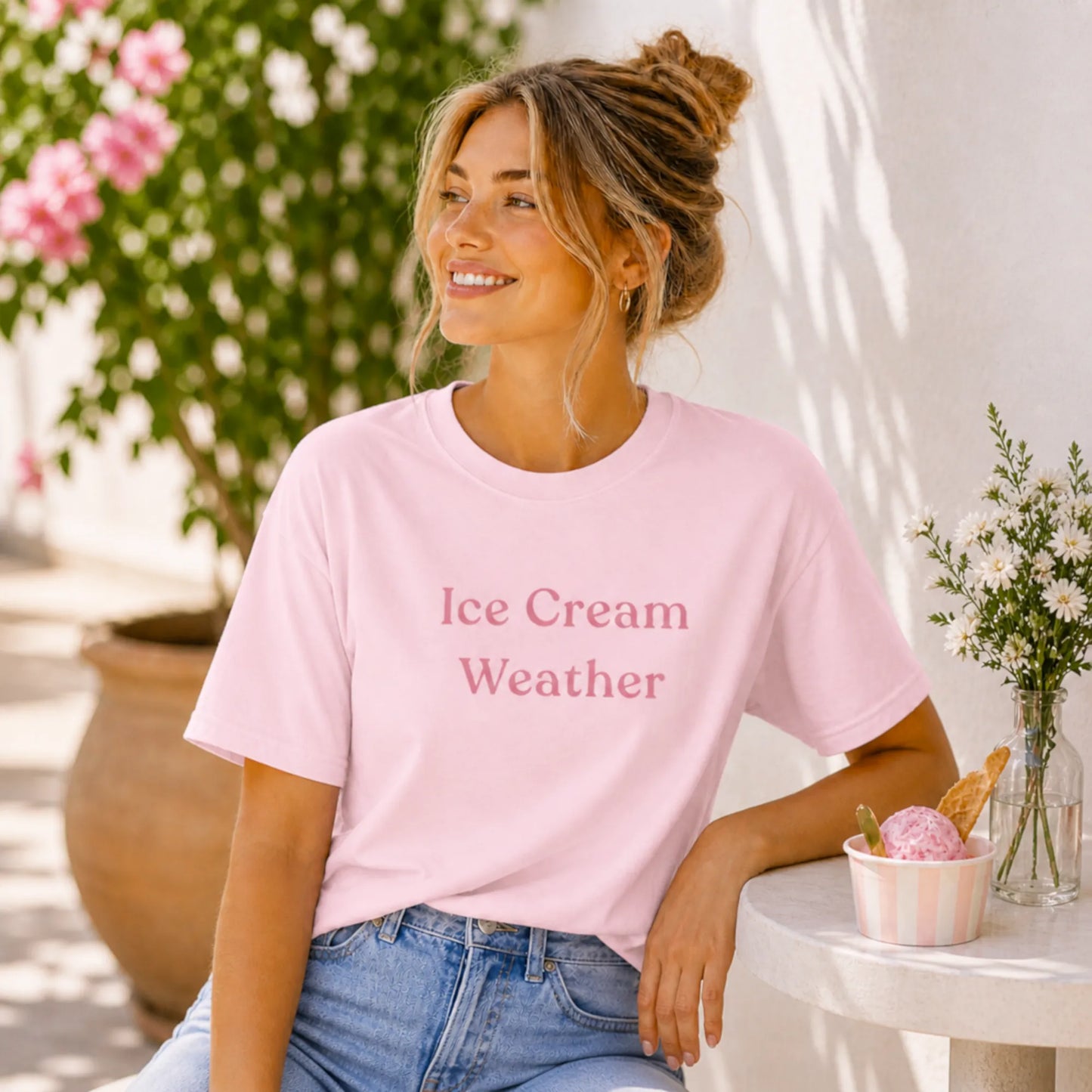 Female model wearing the Ice Cream Weather T-Shirt in soft pink, sitting outdoors in a sunny summer setting with flowers and ice cream.