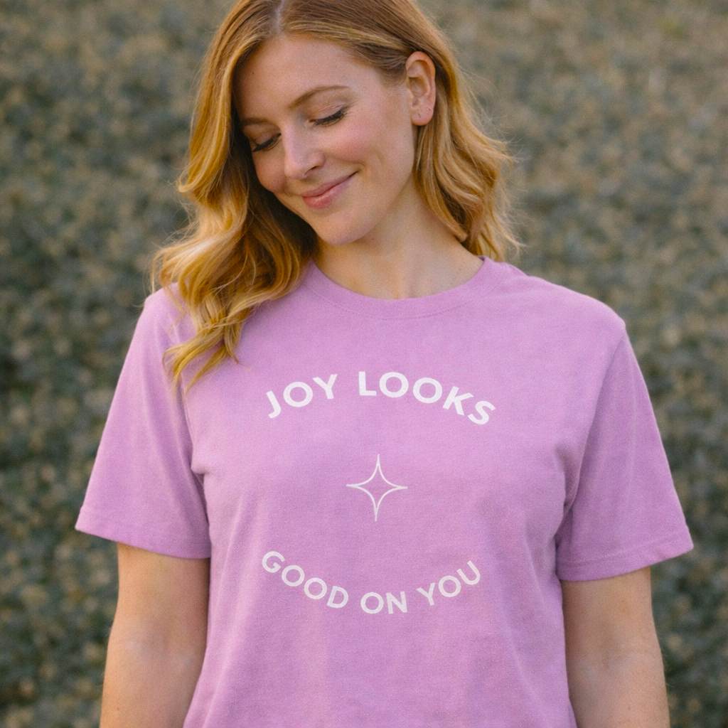 Woman wearing soft lilac “Joy Looks Good on You” graphic T-shirt outdoors in warm natural light with a relaxed, gentle expression