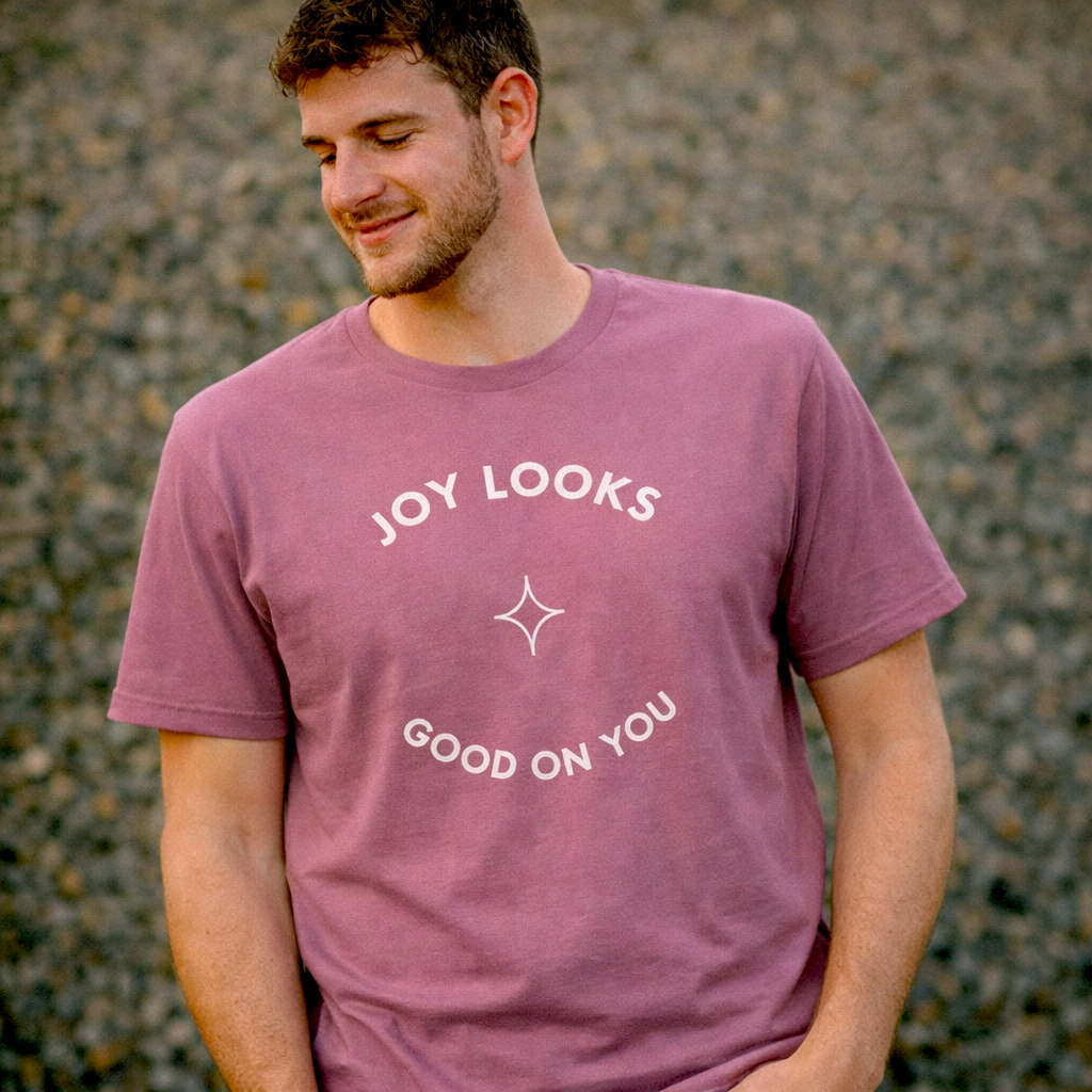 Man wearing muted mauve “Joy Looks Good on You” graphic T-shirt outdoors in soft natural light with a calm, relaxed expression