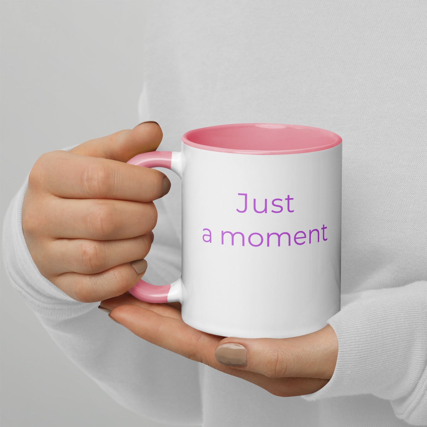 Ceramic coffee mug with pink handle and the words “just a moment”, held in hands with soft knit sleeves, capturing a calm and reflective pause.