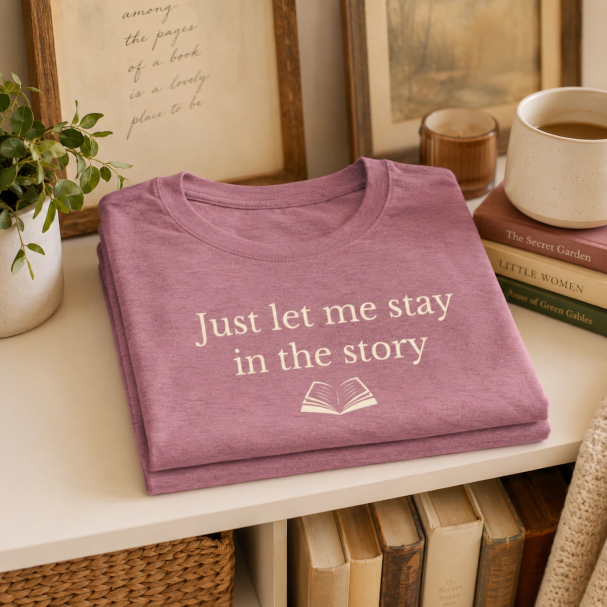 Folded Mauve “Just Let Me Stay in the Story” T-Shirt styled on a shelf with books, a mug, plants and soft reading-inspired home décor.