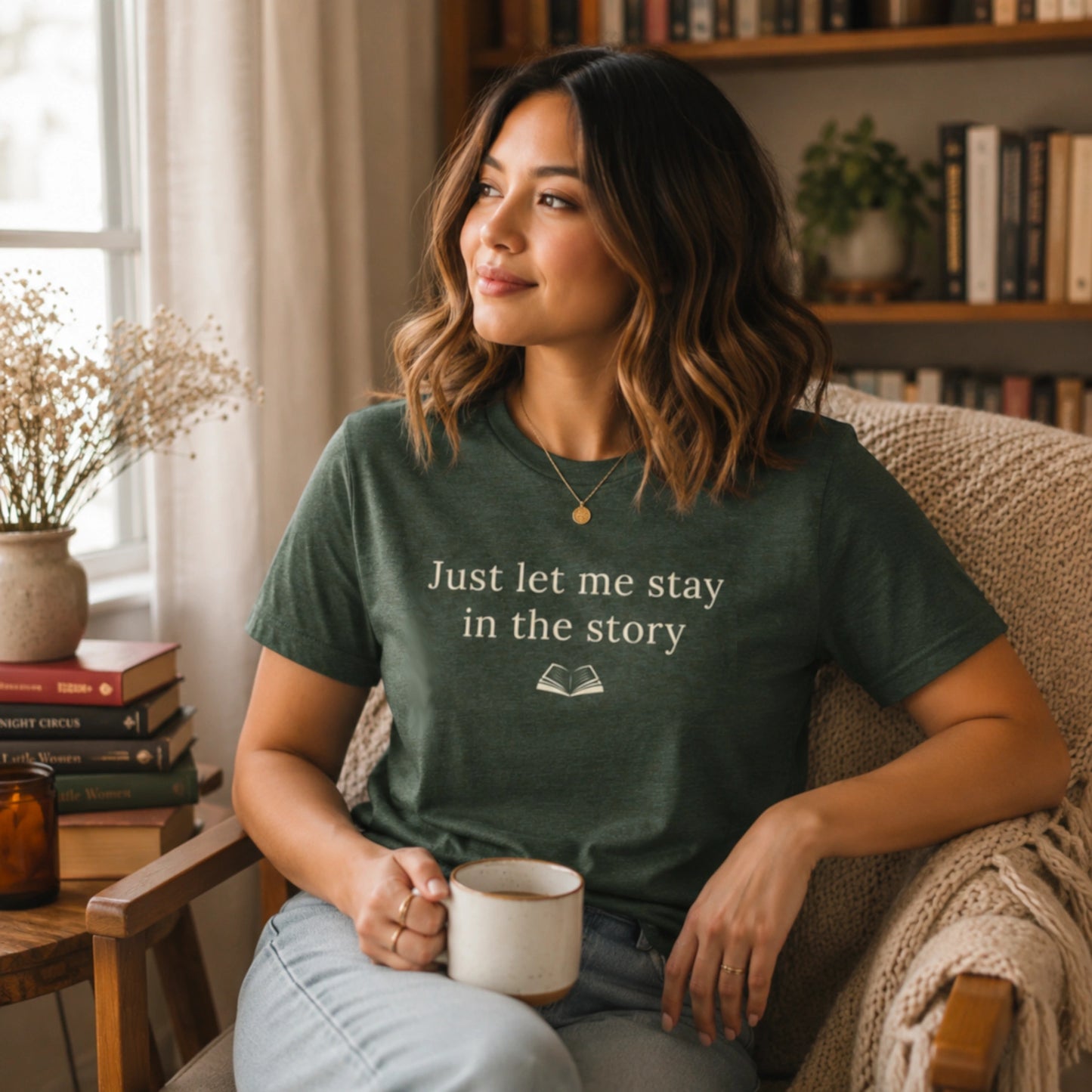 Female model wearing the Heather Forest “Just Let Me Stay in the Story” T-Shirt in a cosy reading nook with books, a mug, soft natural light and warm home details.