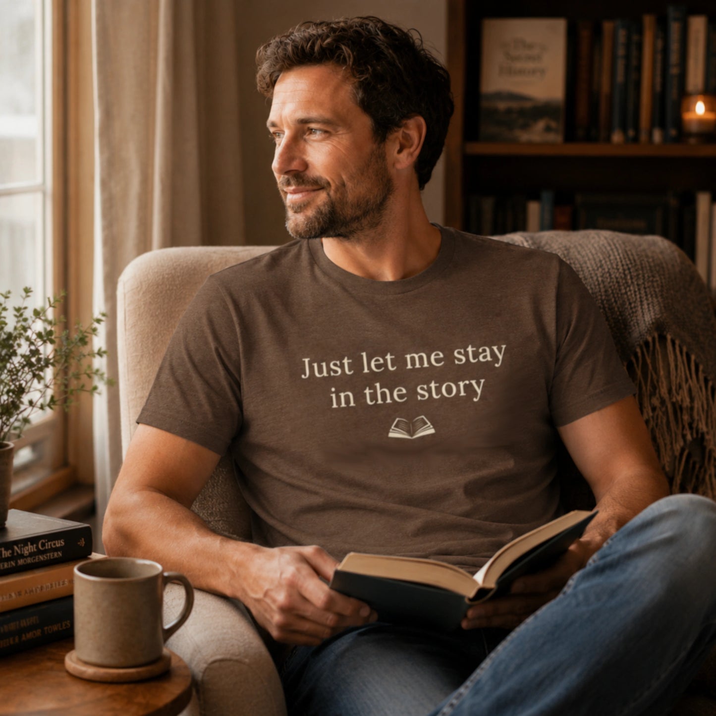 Male model wearing the Heather Brown “Just Let Me Stay in the Story” T-Shirt while reading in a cosy armchair beside books, a mug and warm ambient lighting.