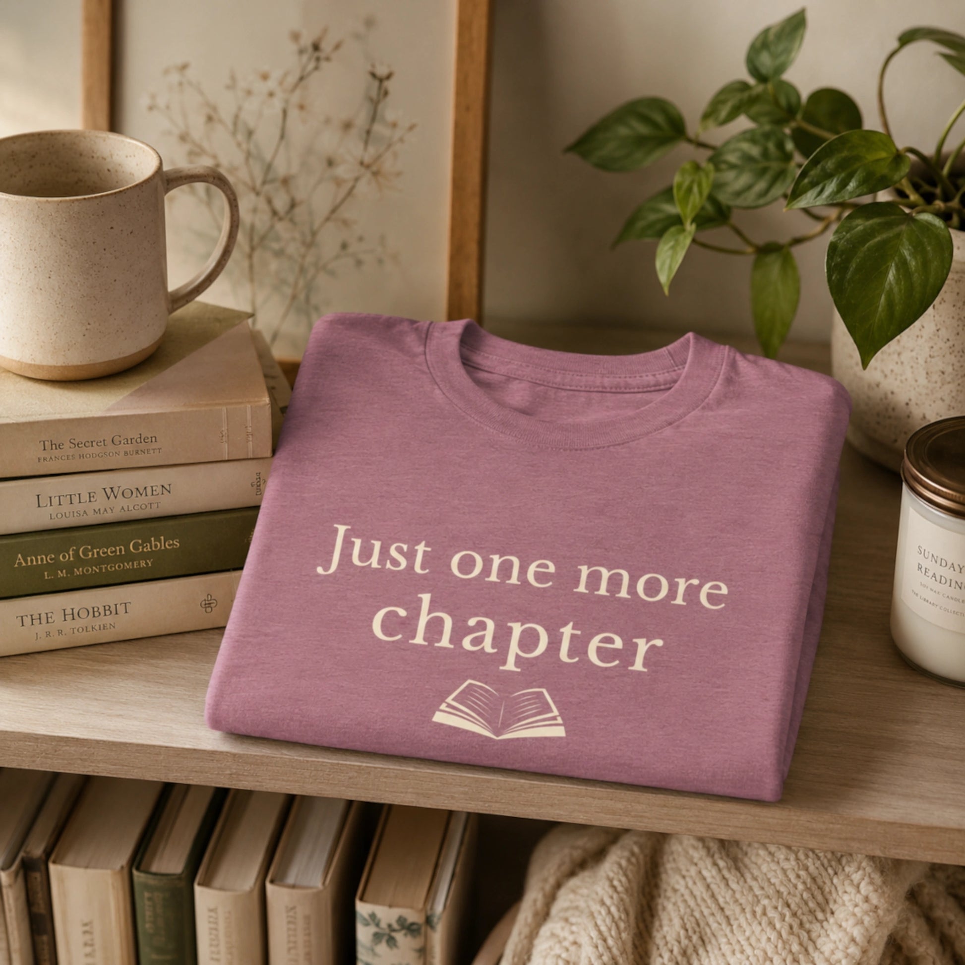 Folded Heather Orchid “Just One More Chapter” T-Shirt styled on a wooden shelf beside books, a mug, a candle and soft home décor.
