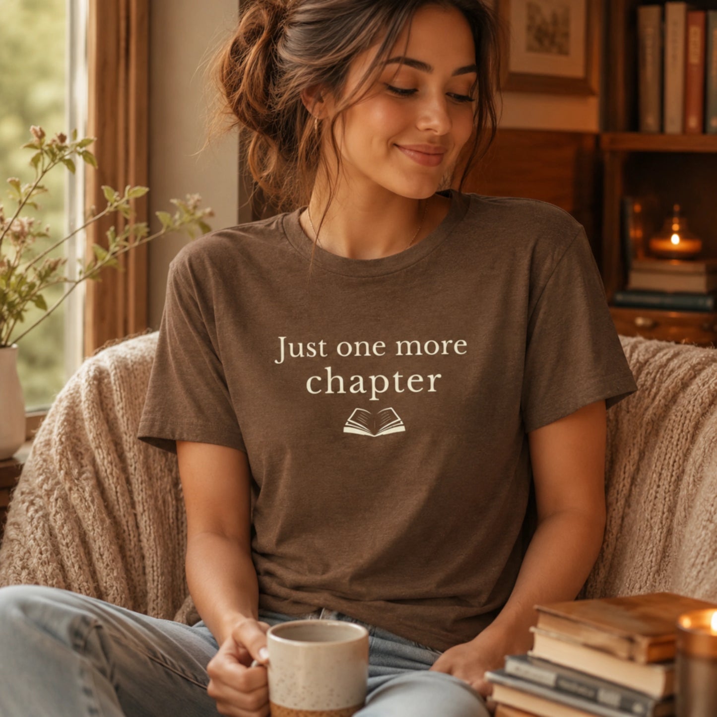 Female model wearing the Heather Brown “Just One More Chapter” T-Shirt in a warm, cosy reading nook with books, a mug and soft candlelight nearby.