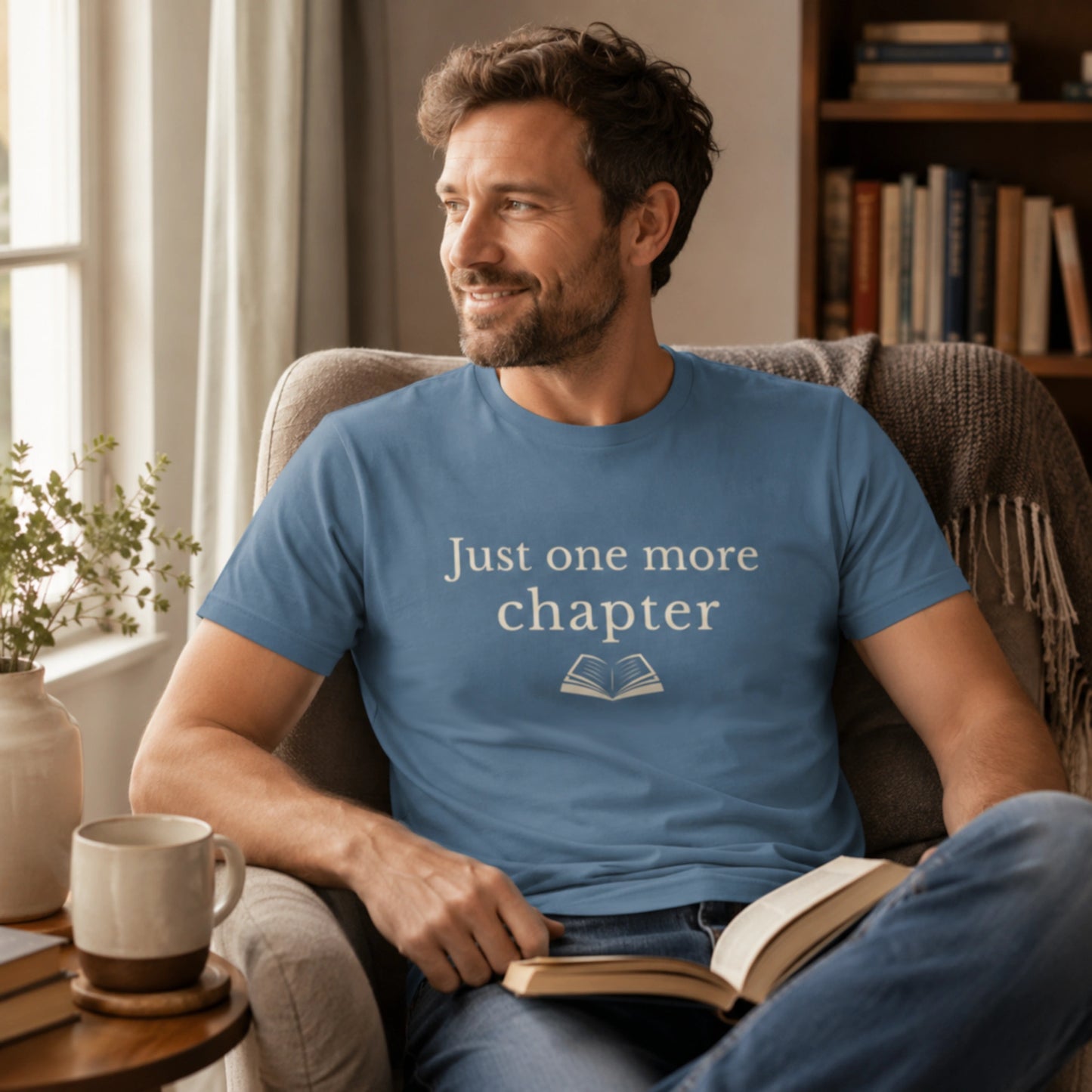 Male model wearing the Steel Blue “Just One More Chapter” T-Shirt while relaxing in a cosy armchair with a book and mug in a calm reading corner.