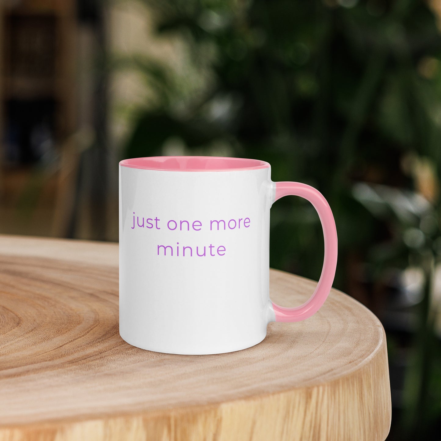 Ceramic coffee mug with pink handle and the words “just one more minute”, placed on a wooden table with warm light and soft textures, creating a peaceful and relaxed morning setting.