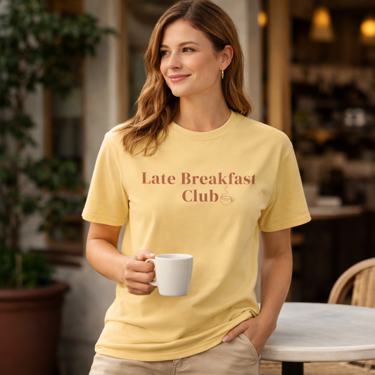 Female model wearing Late Breakfast Club Comfort Colors T-shirt in butter yellow holding a coffee at a café