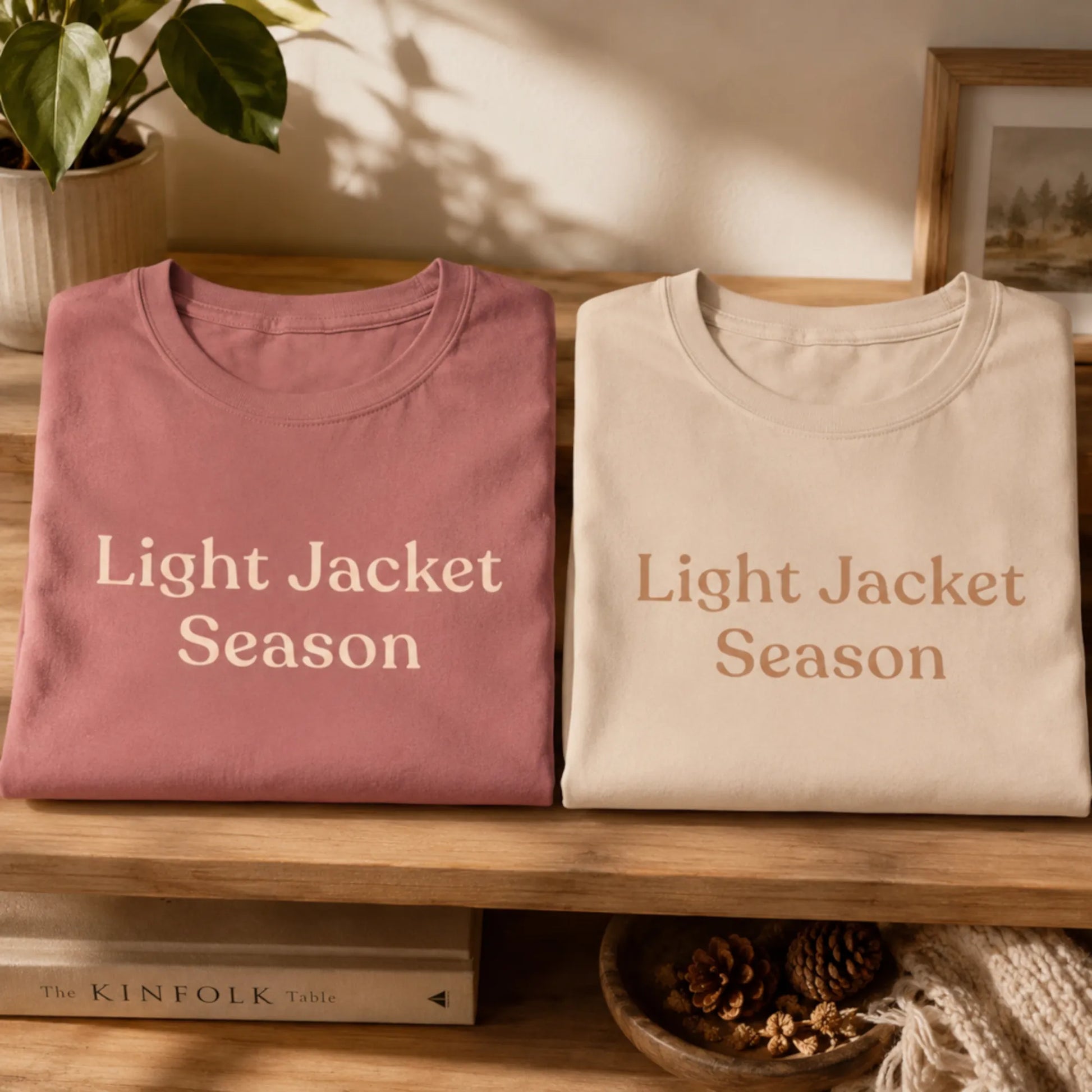 Folded Light Jacket Season T-shirts in mauve and soft cream displayed on a wooden shelf with warm natural light and cosy seasonal styling.