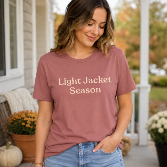 Woman wearing the Light Jacket Season T-shirt in mauve with soft cream text, standing outdoors on a porch with a calm autumn feel.