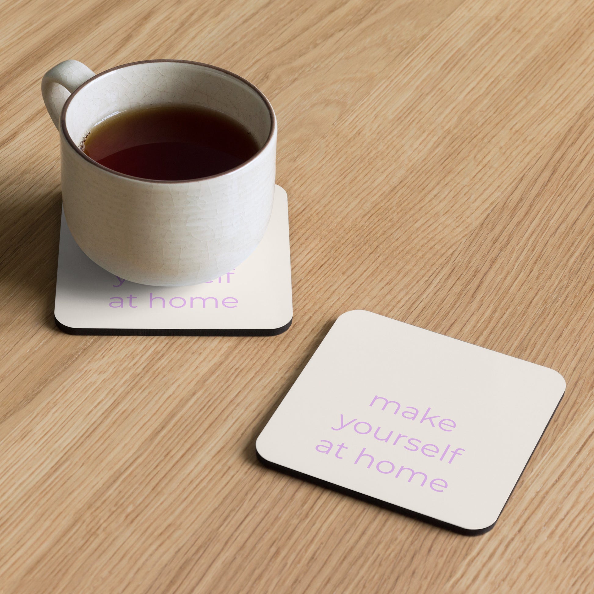 Neutral coaster with “make yourself at home” text holding a mug on a wooden table in soft natural light