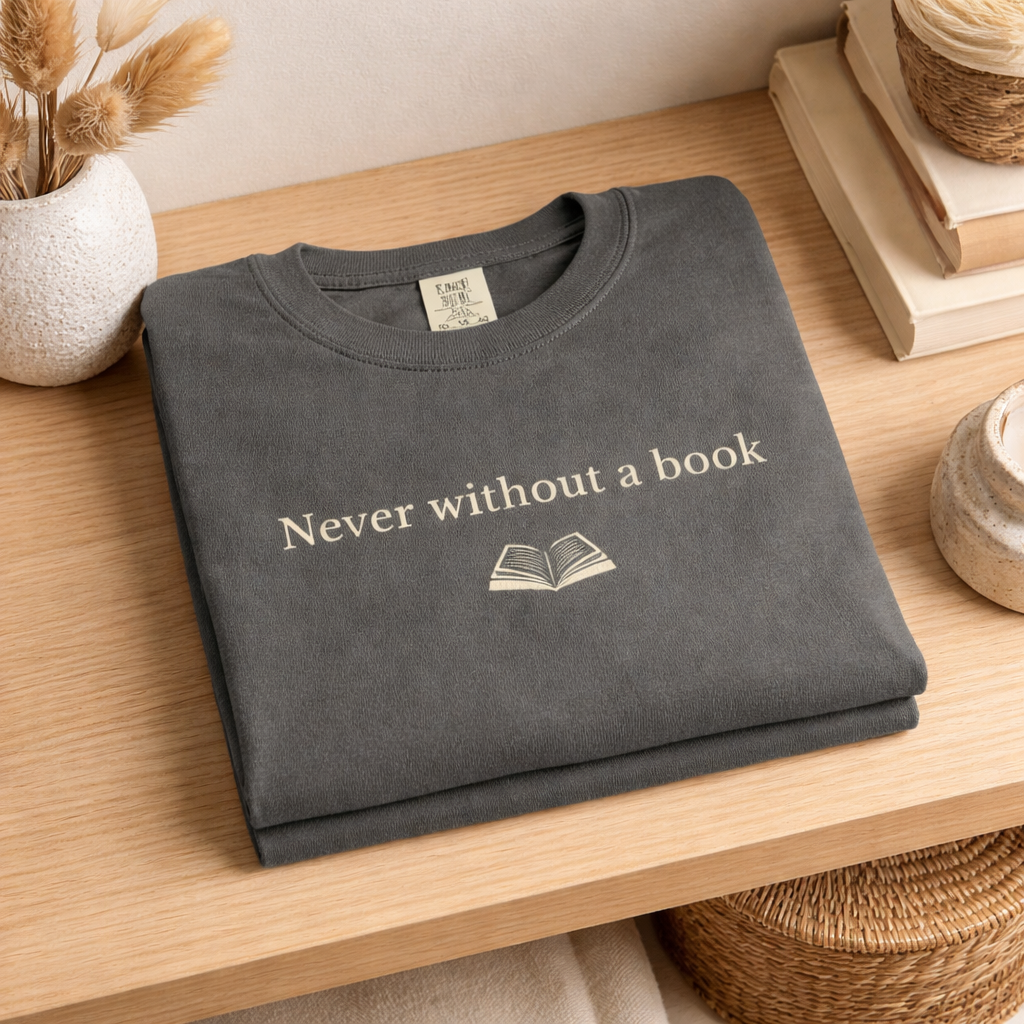 Folded charcoal Comfort Colors T-shirt with “Never without a book” text displayed on a wooden shelf with books and warm neutral decor
