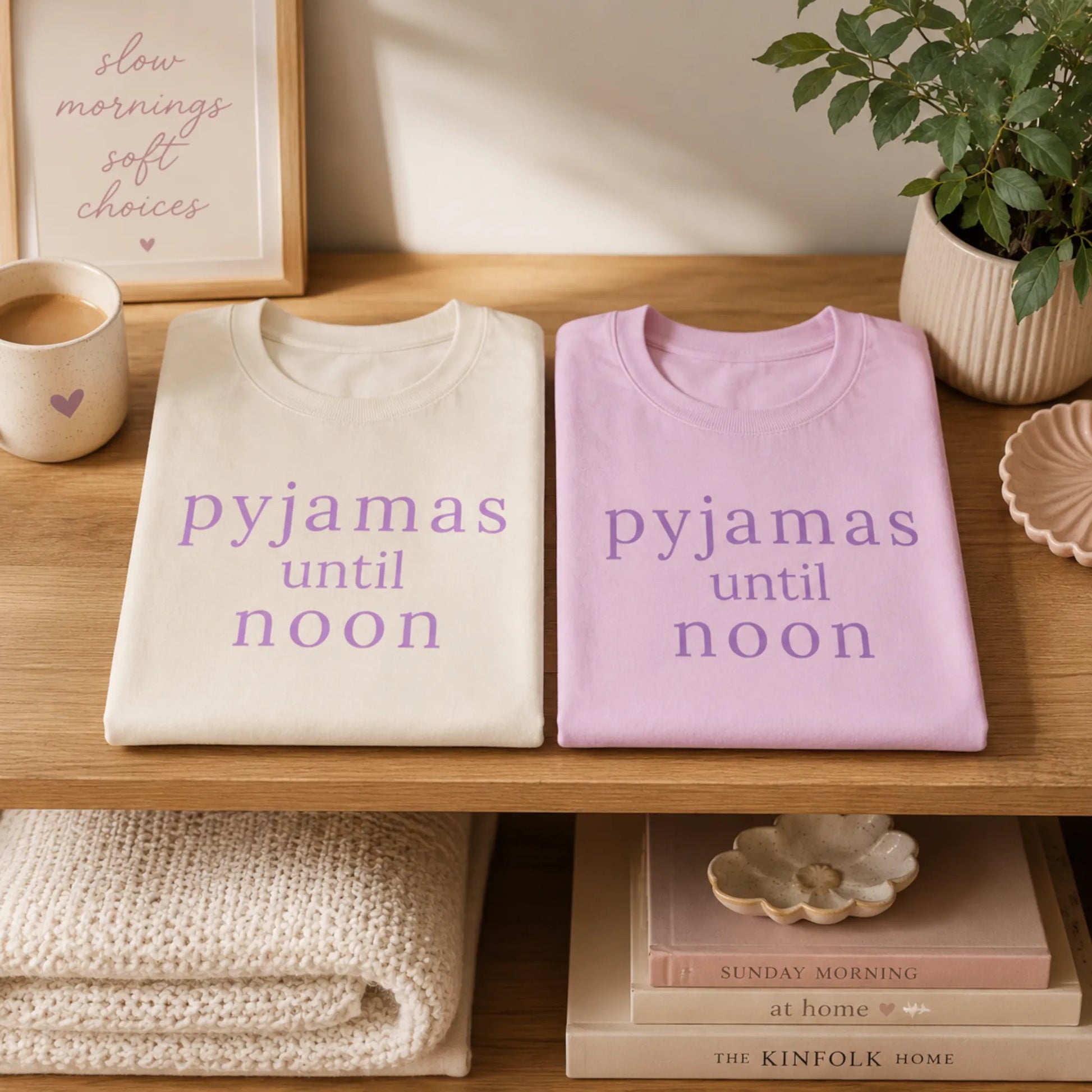 Folded Pyjamas Until Noon T-shirts on a warm wooden shelf in soft natural light, showing the natural and lilac colourways with cosy slow morning styling.