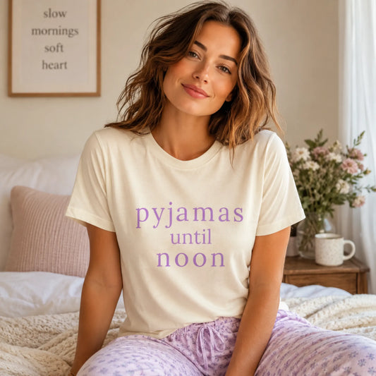 Female model wearing a natural Pyjamas Until Noon T-shirt in a soft cosy bedroom setting, styled for a relaxed slow morning at home.