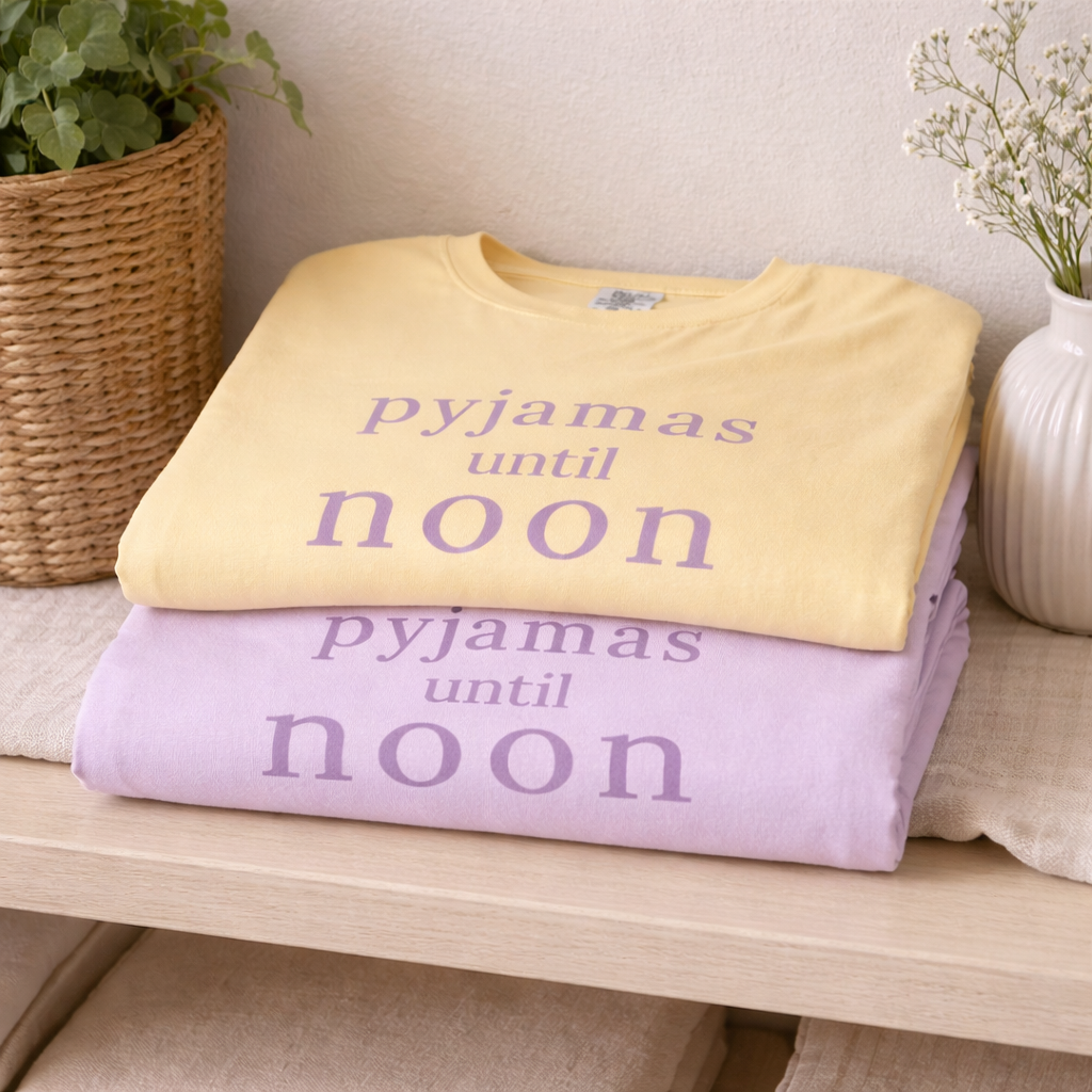 Folded butter and orchid Pyjamas Until Noon Comfort Colors T-shirts styled on a wooden shelf with plants and soft decor