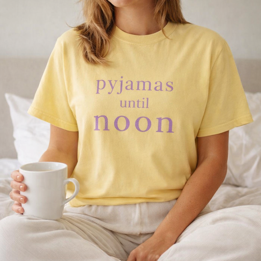 Woman relaxing in bed wearing butter yellow Pyjamas Until Noon Comfort Colors T-shirt holding a mug on a slow Sunday morning