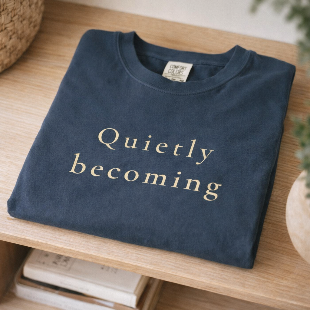 Folded midnight blue Quietly Becoming T-shirt on wooden shelf with soft neutral styling