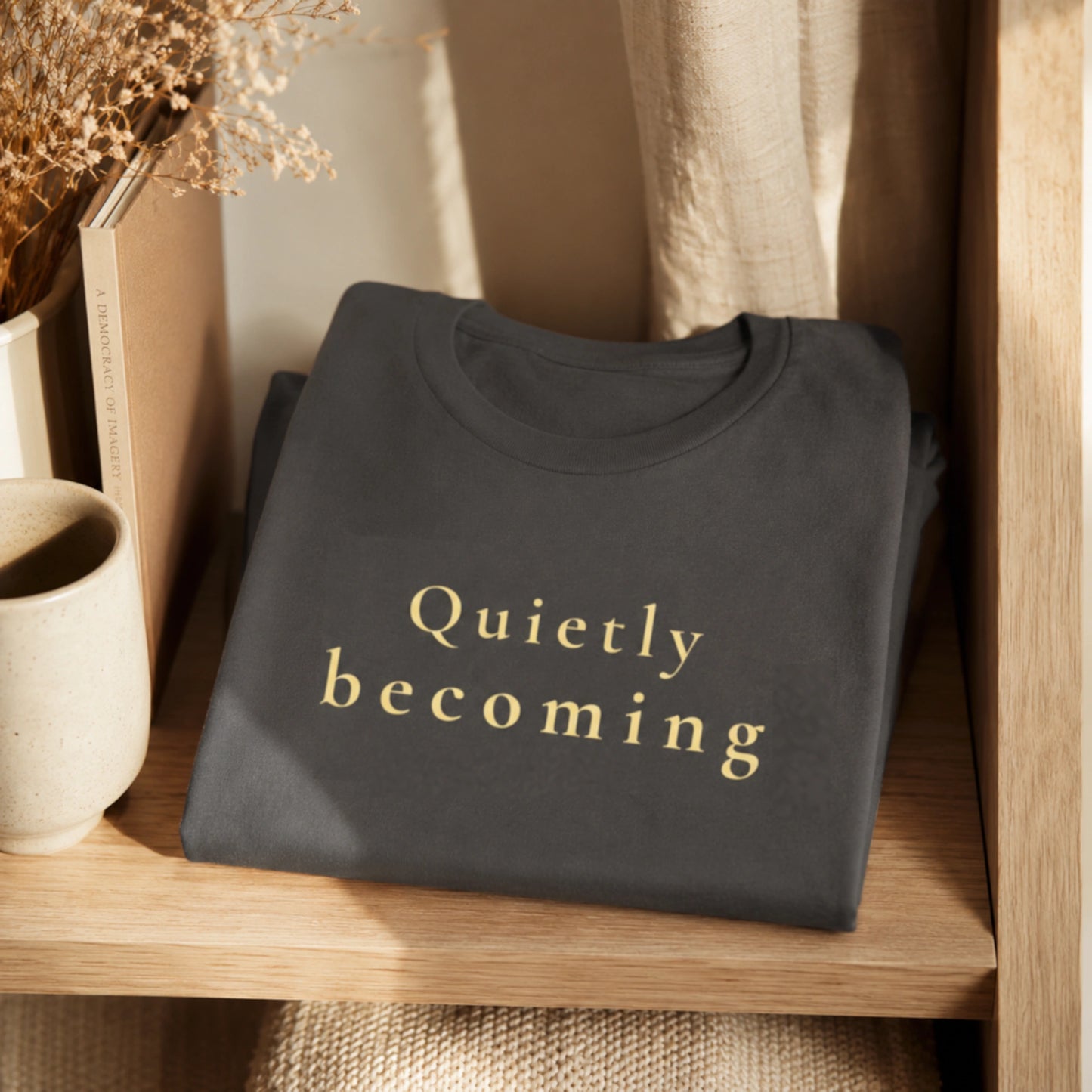 Folded asphalt Quietly Becoming T-shirt displayed on a warm wooden shelf with neutral home styling.