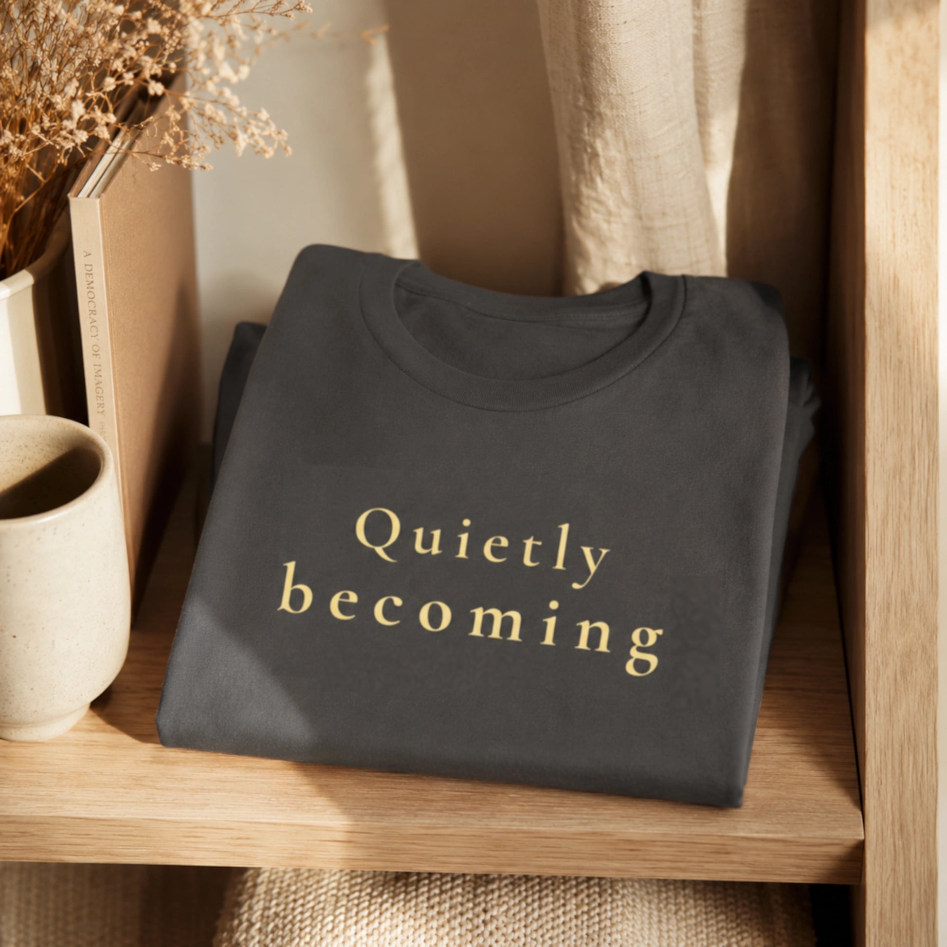 Folded asphalt Quietly Becoming T-shirt displayed on a warm wooden shelf with neutral home styling.