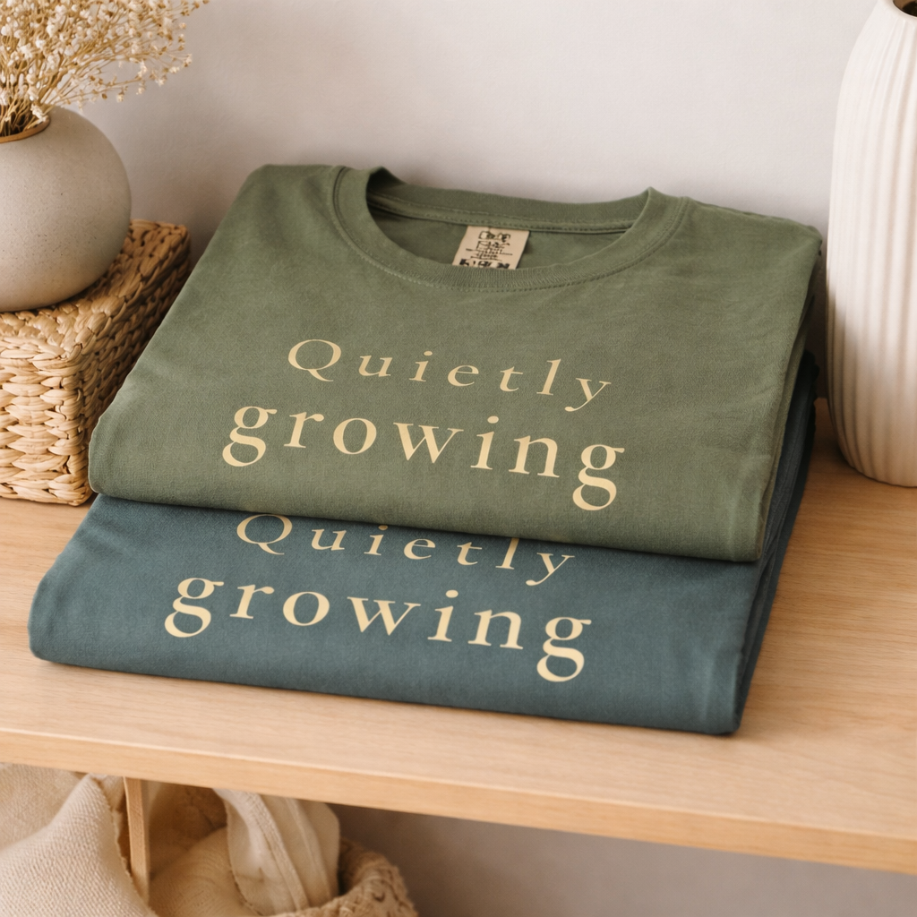 folded sage and blue spruce Comfort Colors T-shirts on a wooden shelf with “Quietly growing” text, warm neutral home setting